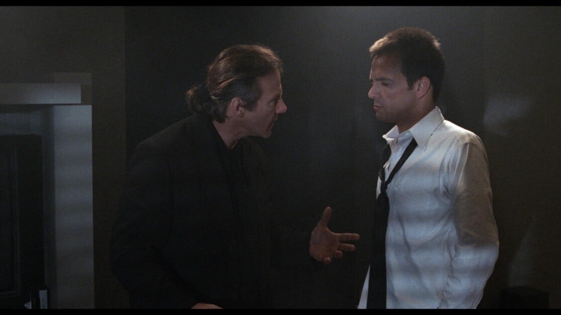Two men in a dimly lit room are having a serious conversation. The man on the left gestures with his hands, while the man on the right, wearing a white shirt and loosened tie, listens intently.