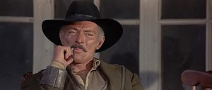 A middle-aged man in a cowboy hat and jacket pensively touches his mustache, sitting by a window.