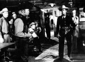 A black-and-white scene from a Western film with men in cowboy attire, one holding a gun, standing in a saloon.
