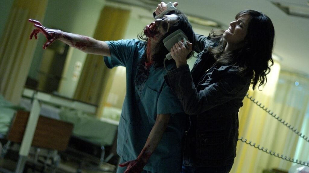 A woman holds a disfigured, zombie-like figure with a medical defibrillator in a dimly lit room, appearing to fend it off.
