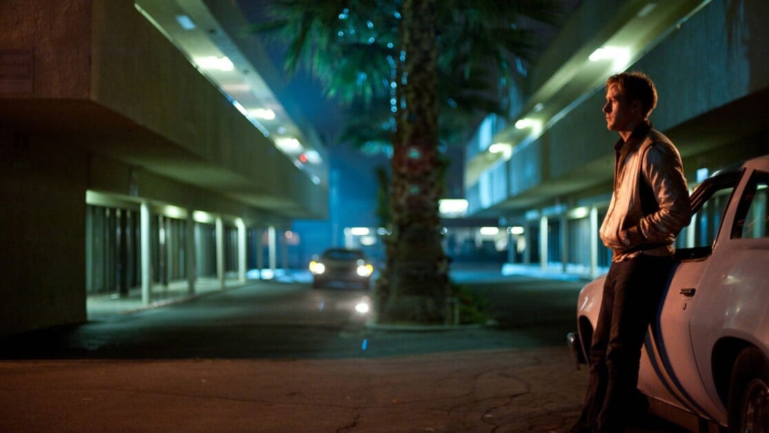 A person in a jacket leans against a car in a dimly lit, palm tree-lined motel parking lot at night.