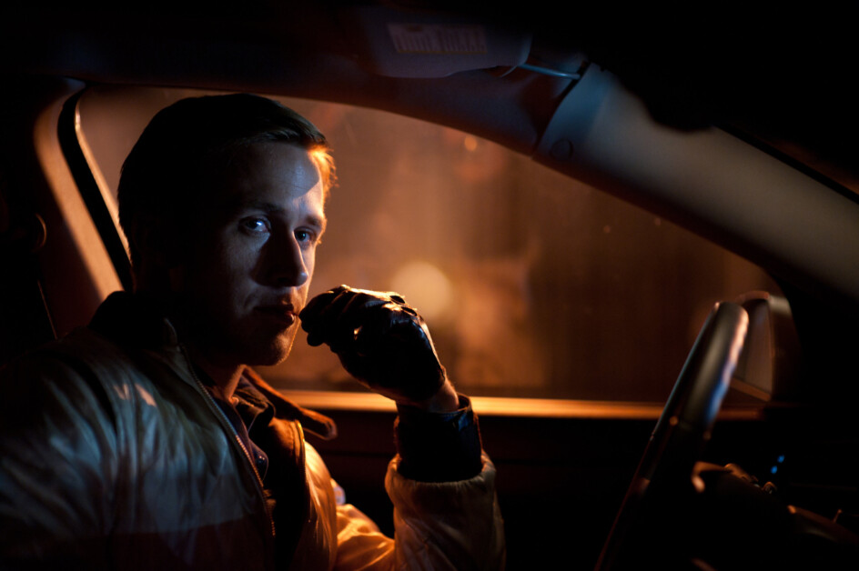 A person in a car at night, wearing a jacket and gloves, holding something near their face while looking towards the camera.