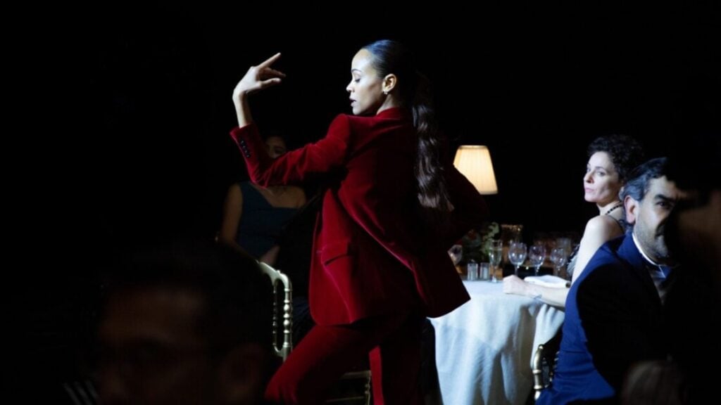 A woman in a red suit stands in a dimly lit room, striking a dramatic pose. Seated guests are visible in the background.
