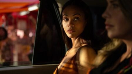 Two women sitting in a vehicle, with one looking at the other who is gazing forward. The background is blurred with red and orange colors visible through the window.