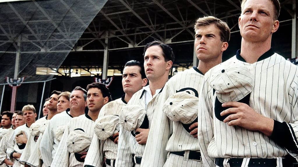 A group of male baseball players in vintage uniforms looking intently to their left in a stadium.