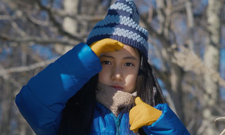 ‘Evil Does Not Exist’ Review – Ryusuke Hamaguchi Makes A Stunning Case For Humanity Young girl in a blue jacket and striped hat touching her forehead, with trees in the background on a sunny day.