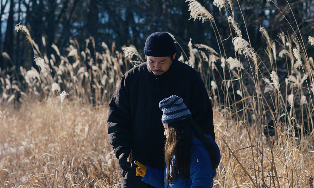 A man in a black jacket and a child in a blue coat walking through a field of tall dry grass in winter.