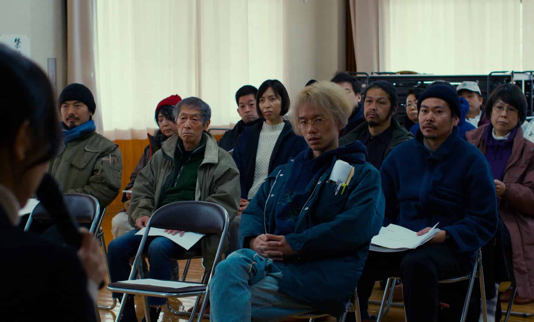 Audience of asian adults in warm clothing seated indoors listening intently to an unseen speaker.