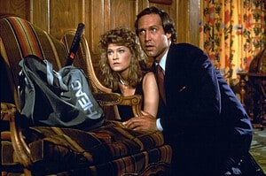 ‘Fletch’ (Special Edition) Blu-Ray Review – Chevy Chase Is At His Straight-Faced Best A man and a woman hiding behind a couch in a vintage-styled room, looking anxious and cautious.