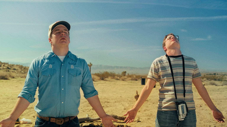 Two men in a desert landscape, one wearing a blue shirt and a cap, the other in a striped shirt and glasses, both looking upwards with bewildered expressions.