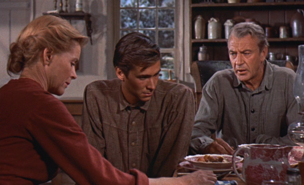 Three people are seated at a wooden table in a rustic kitchen setting, engaged in conversation. Various kitchenware and a window with a view of outside greenery are visible in the background.