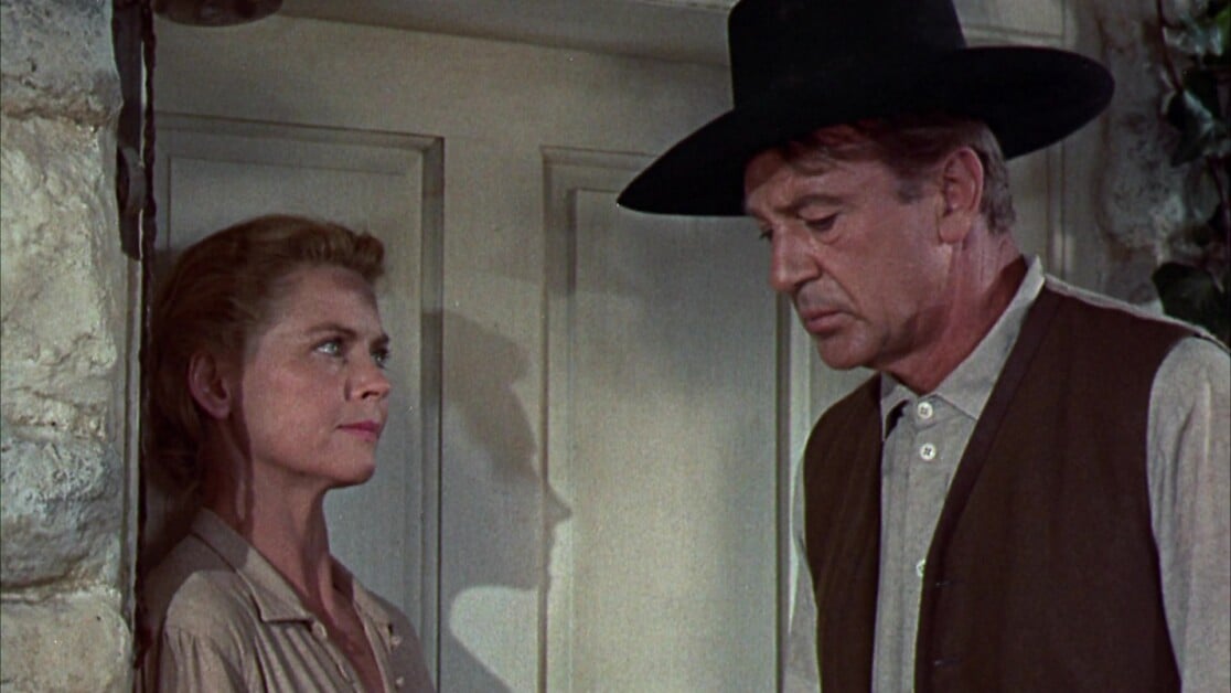 A woman and a man with a black hat stand face to face in front of a door, engaged in a serious conversation.