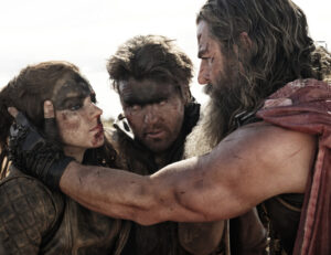 Three people dressed in rugged, medieval-style clothing and with dirt on their faces look intensely at each other. One person is holding the face of another in a serious, concerned manner.
