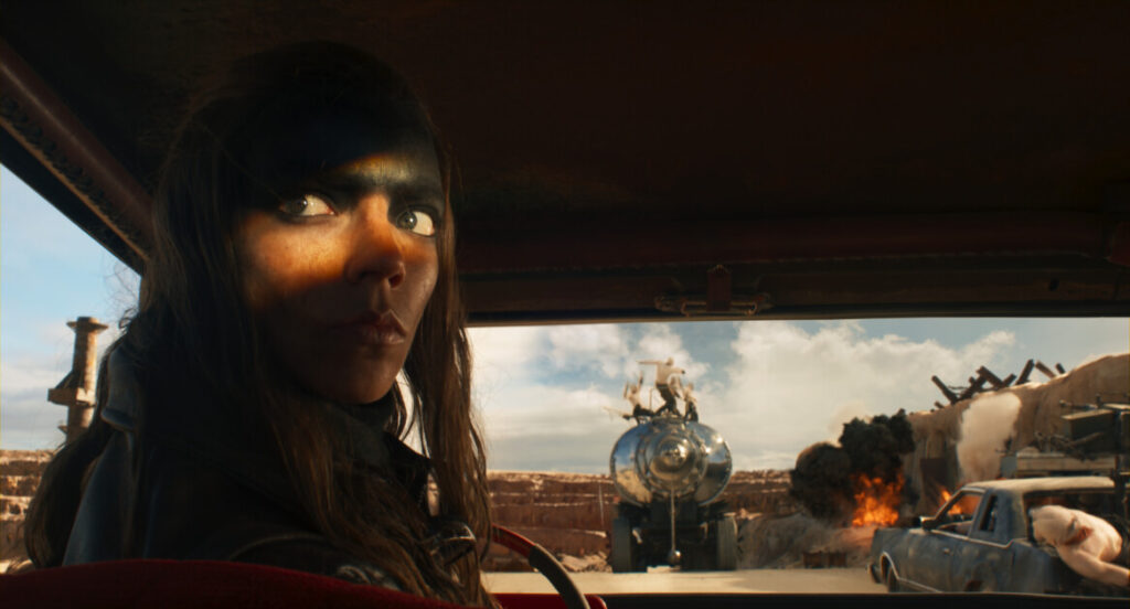 A woman drives a vehicle through a desolate area, with a large spherical machine and a burning car visible through the windshield.