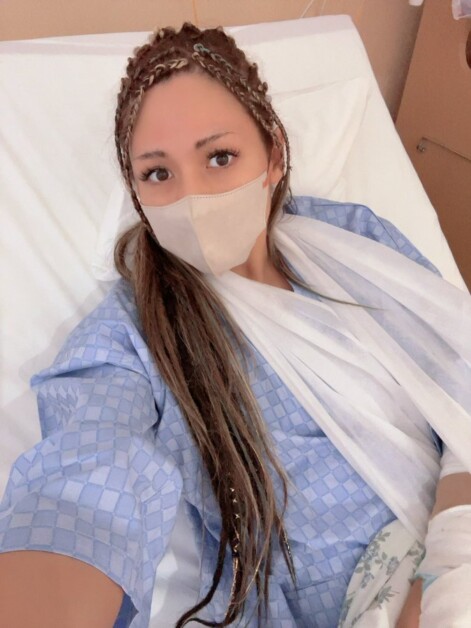 A woman wearing a face mask and a hospital gown is lying in a hospital bed with her arm in a sling.
