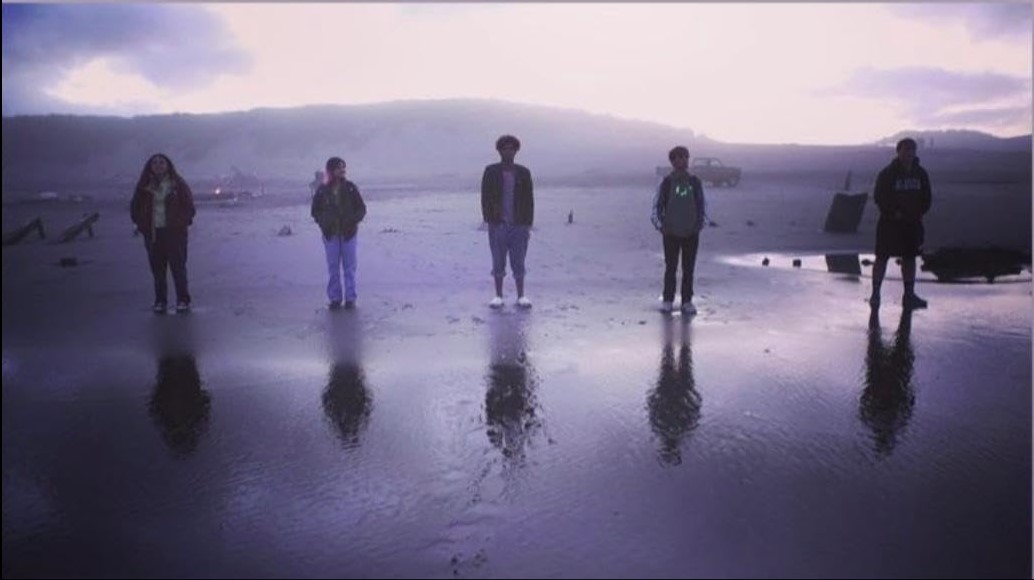 Five people are standing on a wet, reflective beach with a foggy background and hills in the distance.