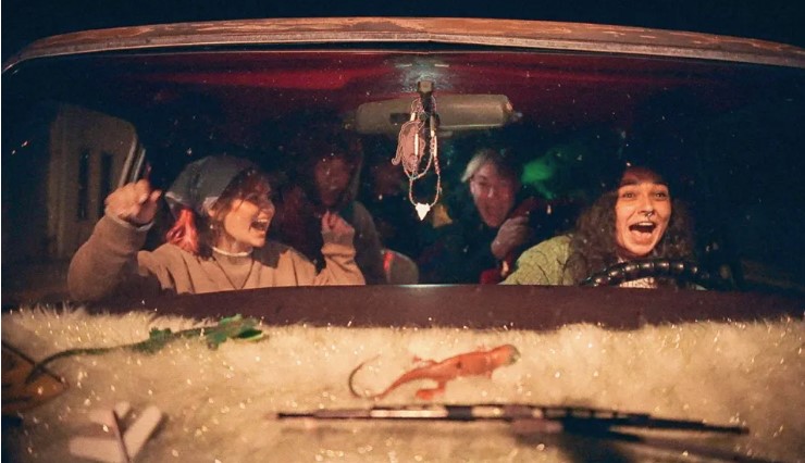 Four people inside a car at night. The driver and front passenger are smiling and appear excited, while the two rear passengers are cheering. Decorations are visible on the car's dashboard.