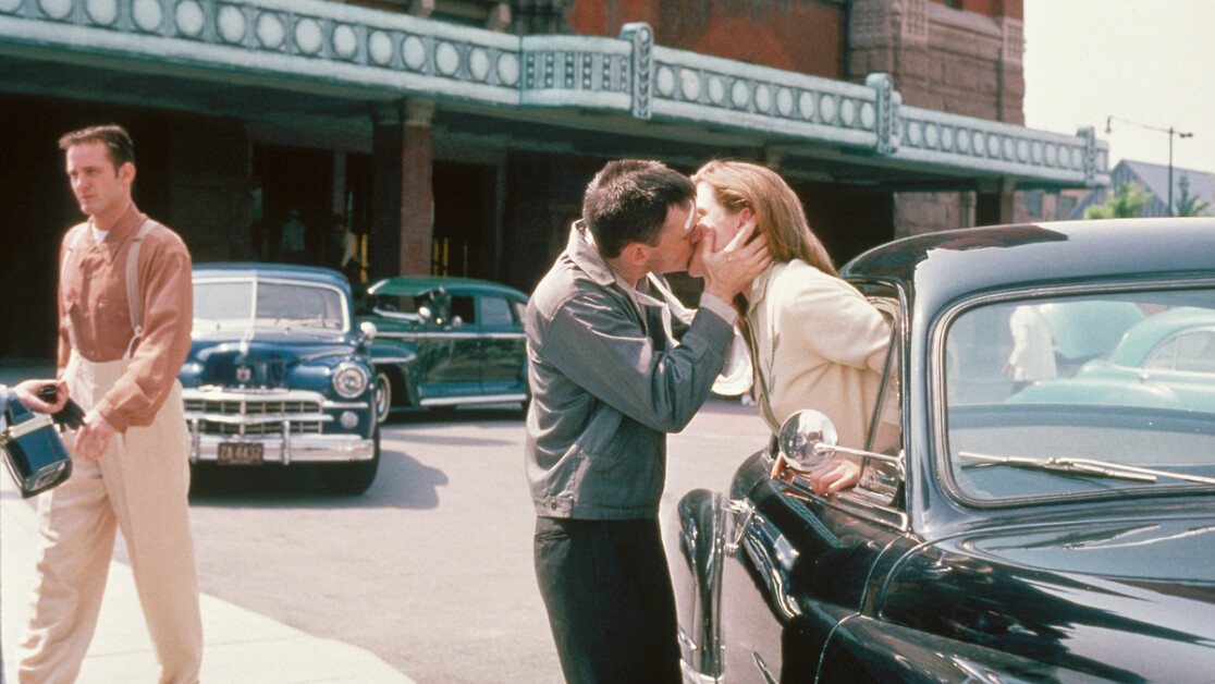 A man and woman kiss through the window of a black car parked on a city street, while another man walks by holding a camera.