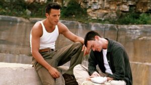 ‘Going All The Way: The Director’s Edit’ Blu-Ray Review – Davies & Affleck Search For Their Place In The World Two people sit on a rock formation. One, in a white tank top, comforts the other, who appears to be in distress and has a bandaged hand. The background shows a rocky cliff.