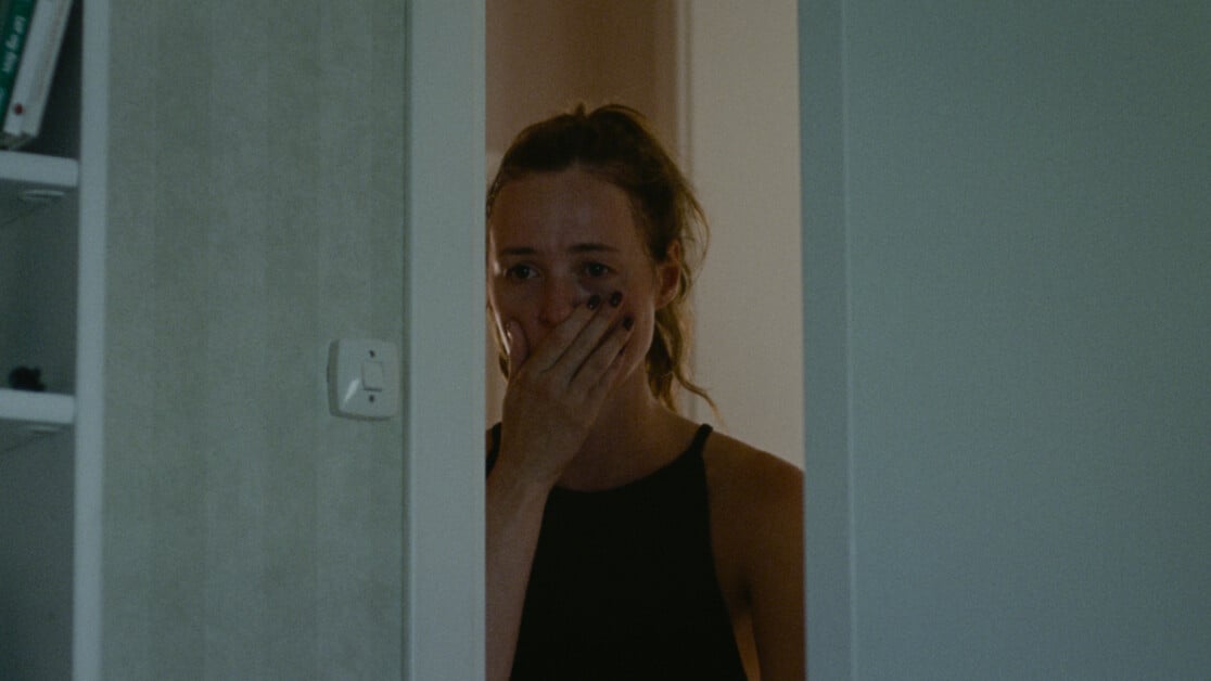 A woman stands in a doorway with her hand covering her mouth, appearing shocked or distressed.