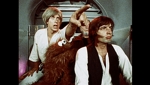 ‘Hardware Wars’ Blu-Ray Review – The Original Star Wars Parody Is Still A DIY Delight Two men and a person in a furry costume inside a spaceship, one pointing urgently while the other looks on, concerned.