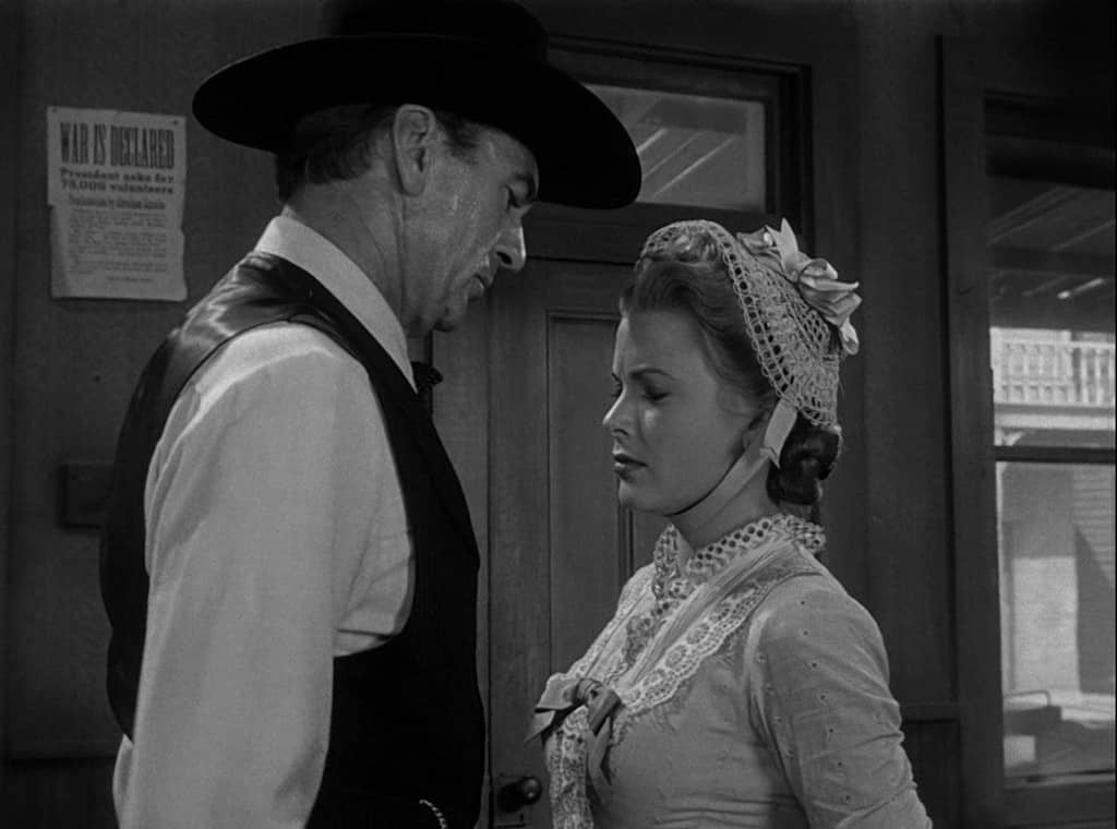 A man in a cowboy hat and suspenders faces a woman wearing a bonnet and lace collar, both looking serious, in a vintage, black-and-white setting.