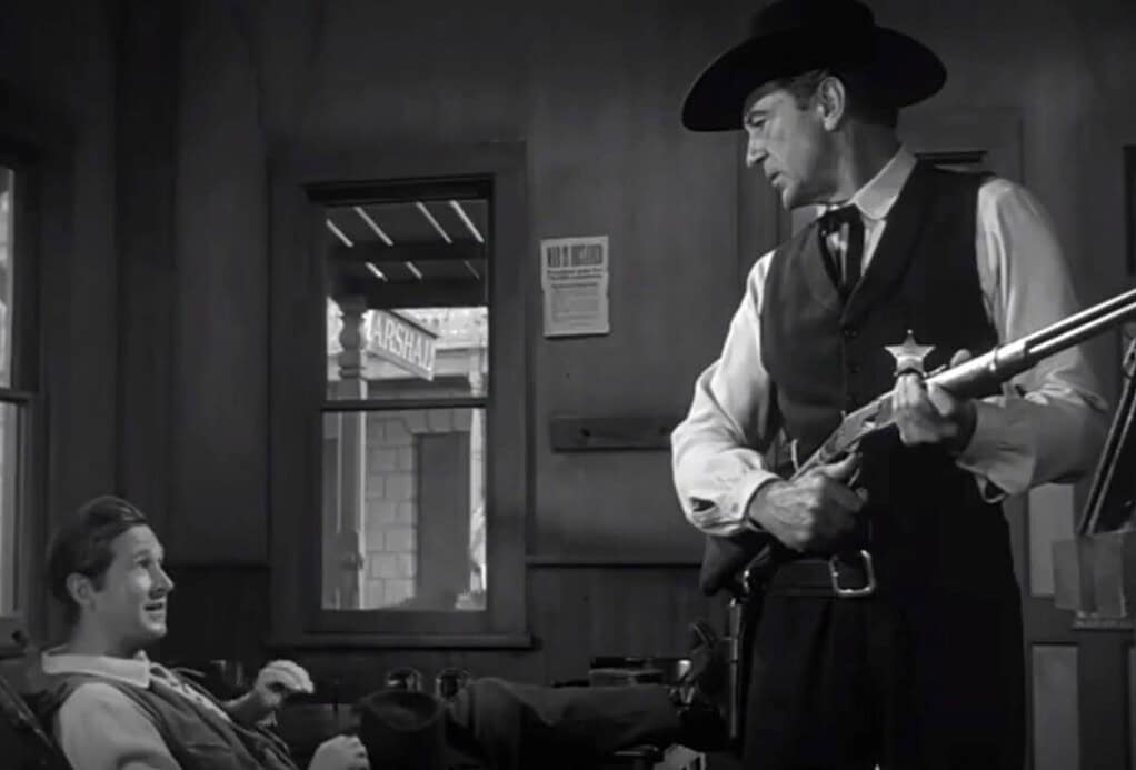 Black and white scene of a sheriff holding a rifle, standing and facing a seated young man in a rustic office setting.