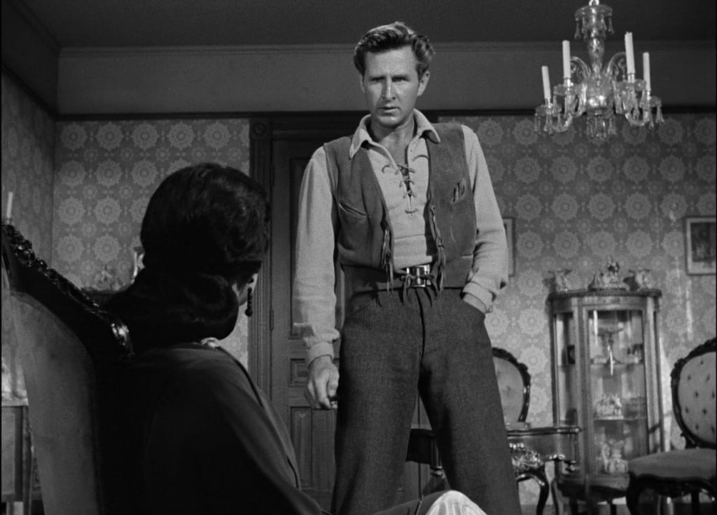 A black-and-white film still showing a man standing and facing a seated person, in a room with vintage decor.