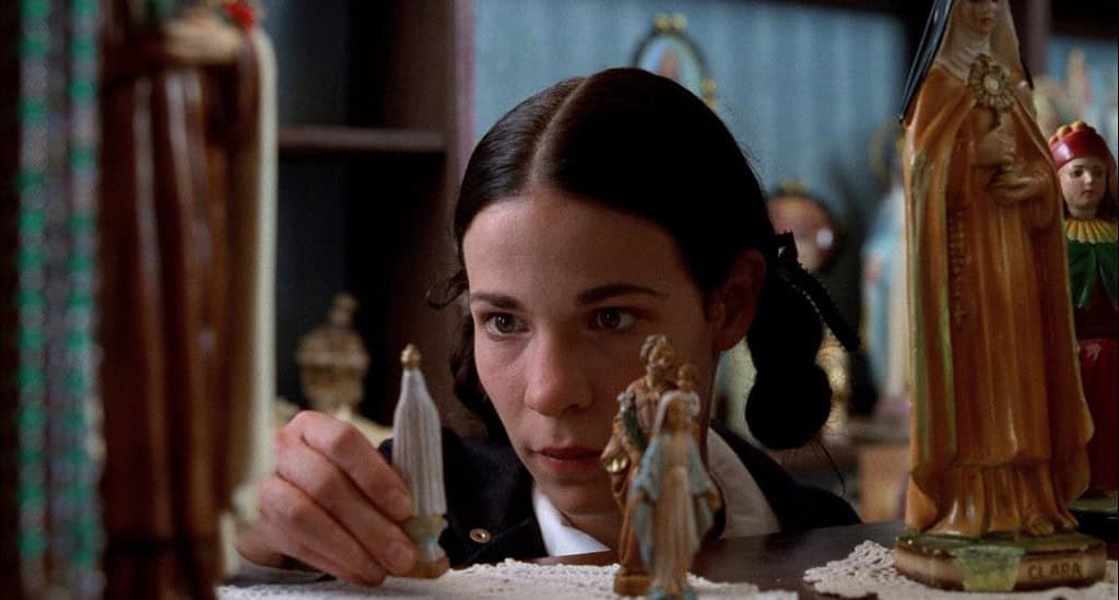 A woman with braided hair examines small religious statues on a table, surrounded by vibrant religious iconography.