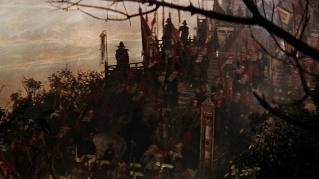A large group of soldiers in traditional armor stands on a fortress wall adorned with flags and banners, surrounded by dense foliage and tree branches.