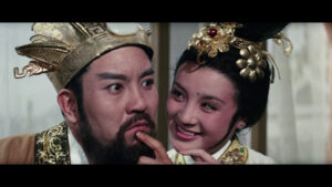 Two actors in traditional Chinese costumes are posing. The man, wearing a decorative headpiece, has a contemplative expression, while the woman smiles while touching his face, also adorned in elaborate attire.