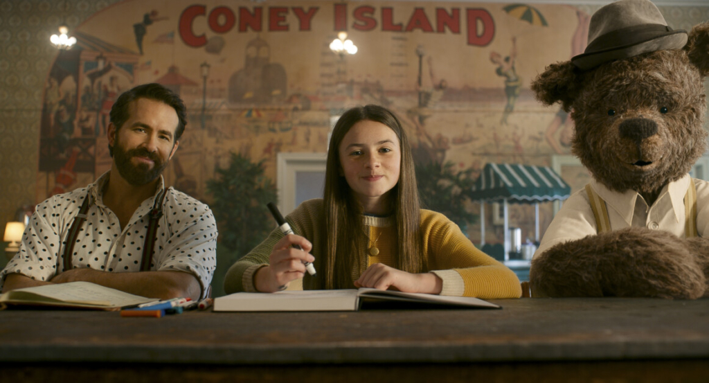A bearded man, a young woman, and a person in a bear costume sit at a table with notebooks in front of them, against a backdrop with "Coney Island" written on it.