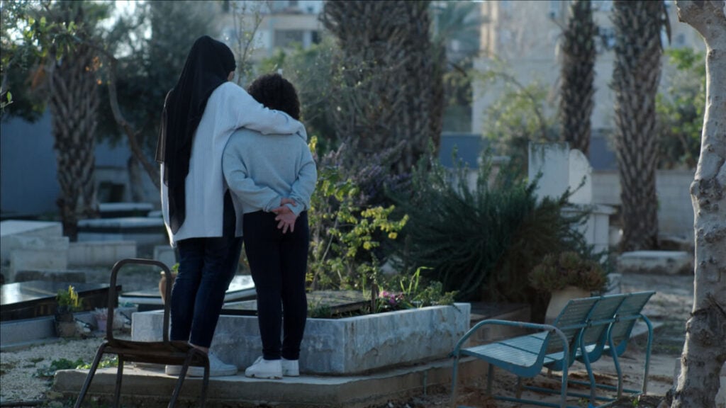 Two people stand together, arms around each other, facing a gravestone in a cemetery with palm trees in the background. A wheelchair and benches are nearby.