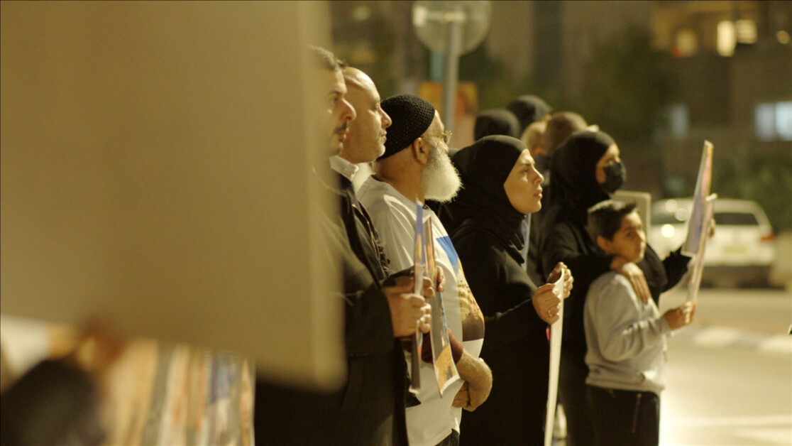 A group of people hold signs during a nighttime gathering on a street, standing in a line and facing forward.