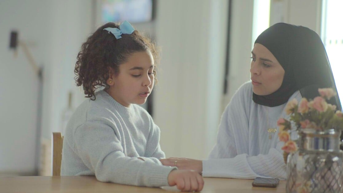 A young girl with curly hair and a blue bow talking to a woman in a hijab at a table with flowers.