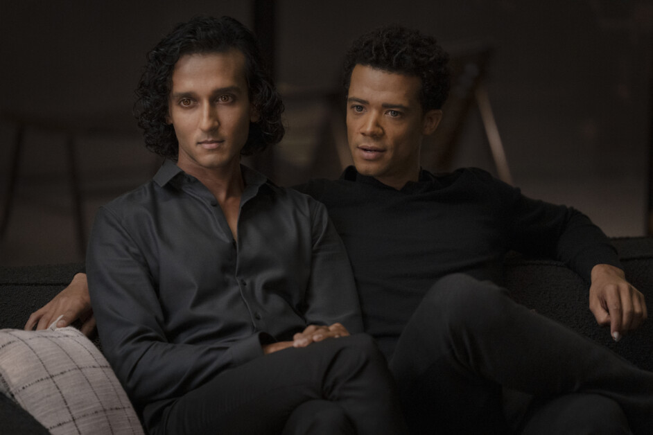 Two individuals with serious expressions sit closely on a dark couch, both dressed in dark clothing. One has curly black hair while the other has short, curly dark hair.