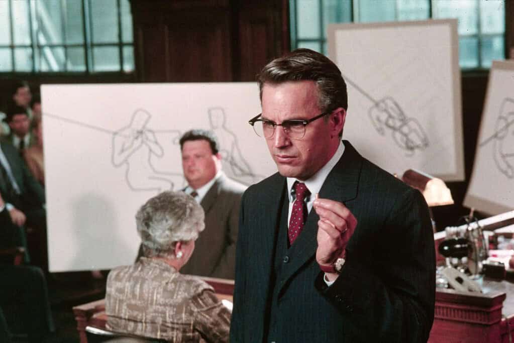 A man in a suit and glasses gestures emphatically in a courtroom setting, with sketches and observers in the background.
