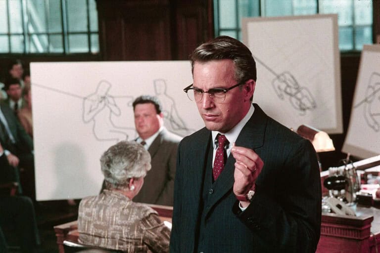 A man in a suit and glasses gestures emphatically in a courtroom setting, with sketches and observers in the background.