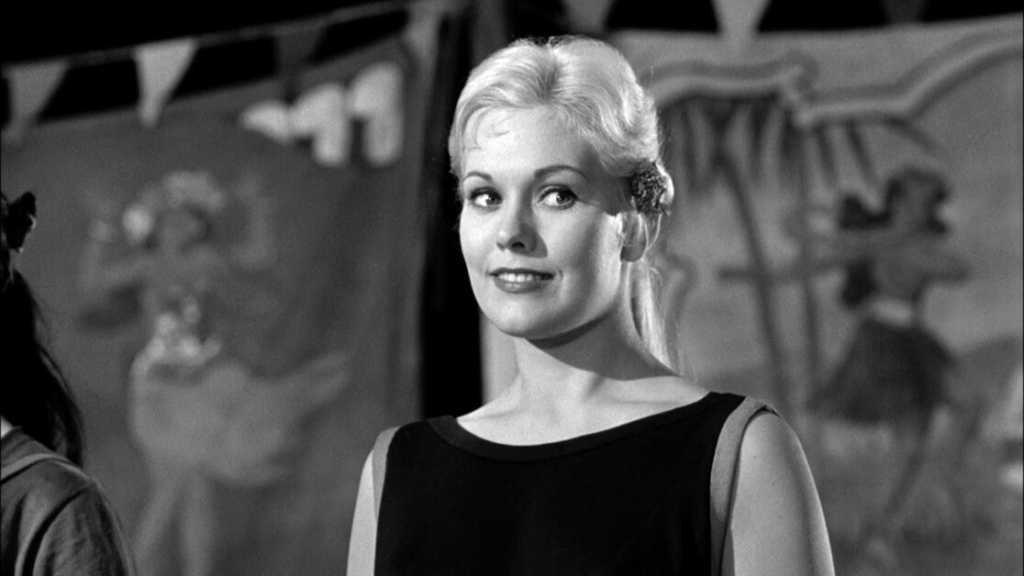 Black-and-white photo of a smiling woman with light hair in a black dress, standing in front of a backdrop with festive decorations.