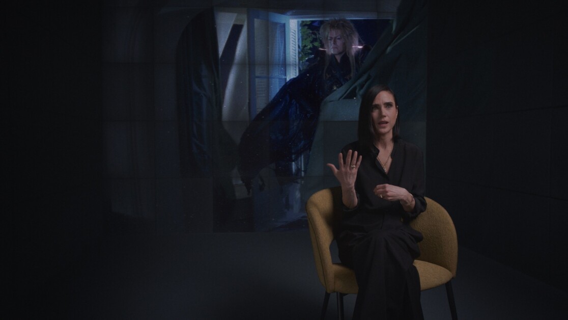 A person sits in a chair gesturing with one hand while a large image of another person appears on the wall behind them.