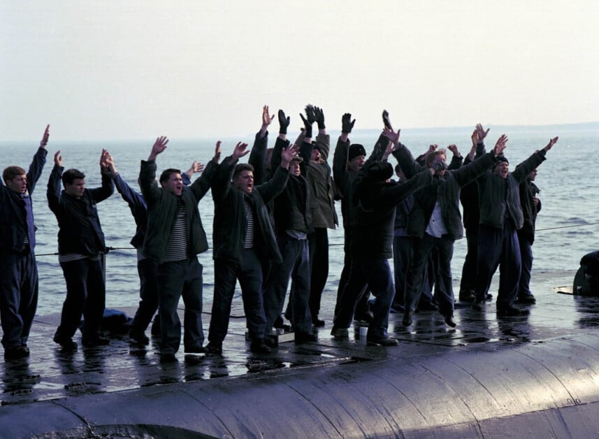 A group of people stands on a submarine, raising their arms while facing the water.
