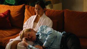 Yorgos Lanthimos Anthology Film ‘Kinds Of Kindness’ Heads To Blu-Ray This October From Searchlight Pictures A woman sits on a couch wearing a white robe, while a man in a blue shirt and tie rests his head on her lap.