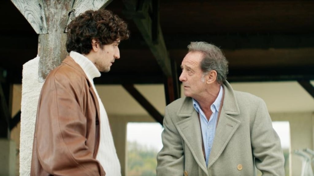 Two men are engaged in a serious conversation outdoors. The man on the left wears a brown leather jacket and a white turtleneck. The man on the right wears a light gray coat over a blue shirt.