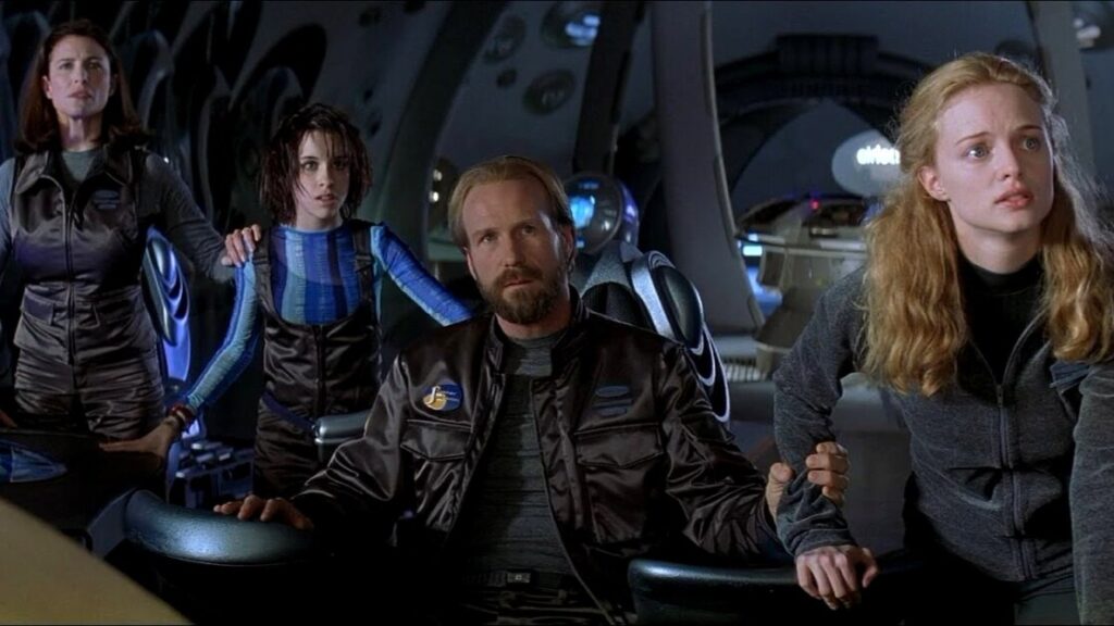 A group of four people in futuristic uniforms stand and sit in what appears to be a spaceship or high-tech control room, looking intently at something out of view.