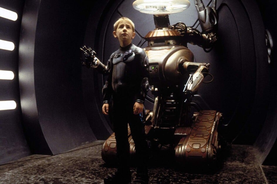A child in a futuristic black outfit stands in front of a large robot with mechanical arms, inside a metallic, dimly lit room.