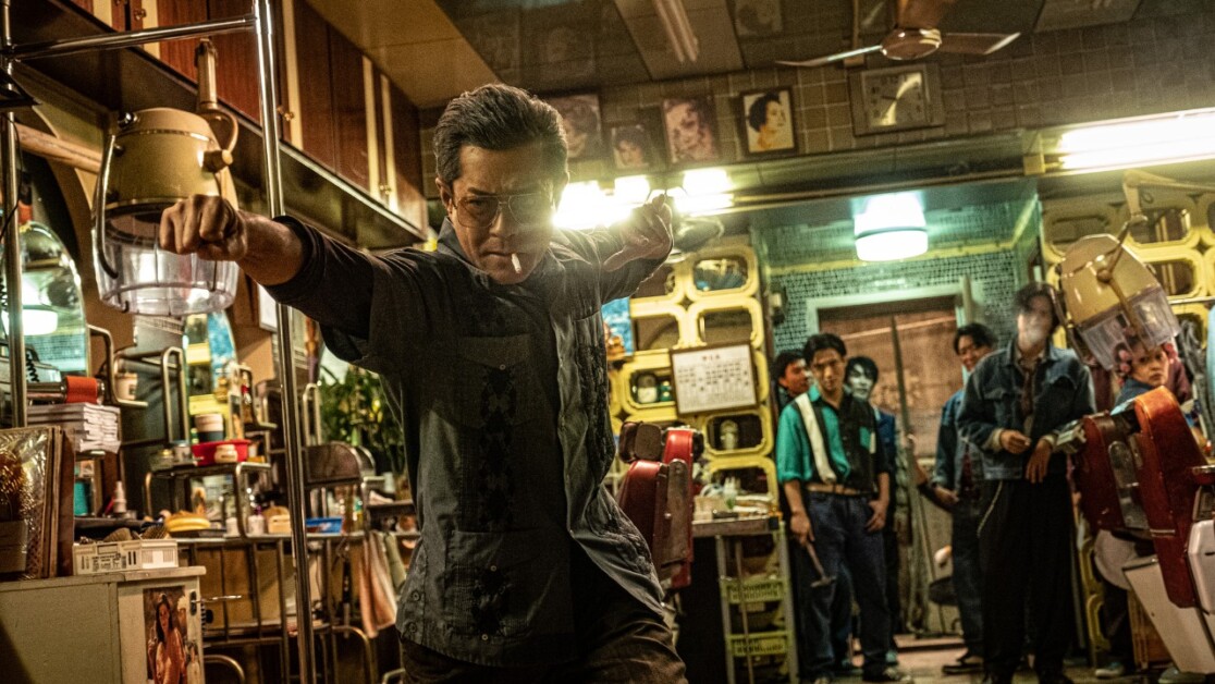 A man in a barber shop poses in a defensive stance with an electric razor, while others in the background observe. The setting is crowded with vintage equipment and decor.
