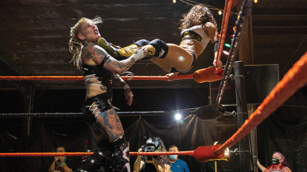 Two female wrestlers in action inside a ring, one kicking the other who is against the ropes.