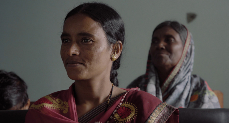 Two Indian women looking towards their left, one in the foreground with a focused expression and another slightly blurred in the background.