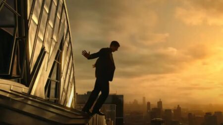 A person in a suit is leaning backward on the edge of a building rooftop at sunset, with a city skyline in the background.