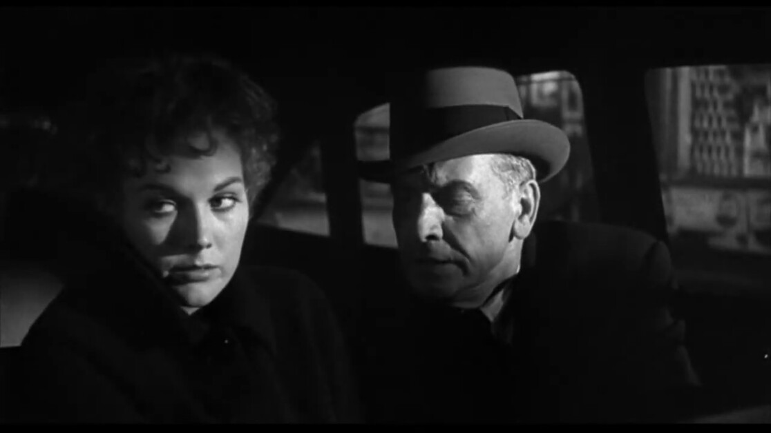 A woman and an older man wearing a hat are seated in a dimly lit car. The woman looks to her left while the man appears to be speaking to her.